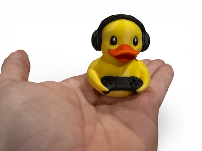 A small yellow duck figurine wearing black headphones and holding a black game controller, displayed on a person's hand against a plain white background.