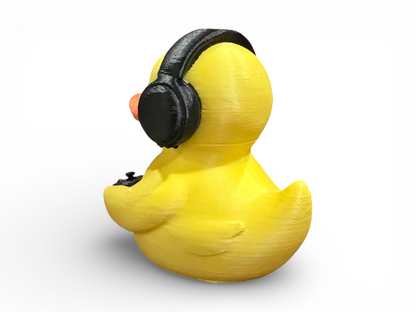 A yellow rubber duck toy shown from the back, wearing black over-ear headphones and holding a black game controller, on a white background.